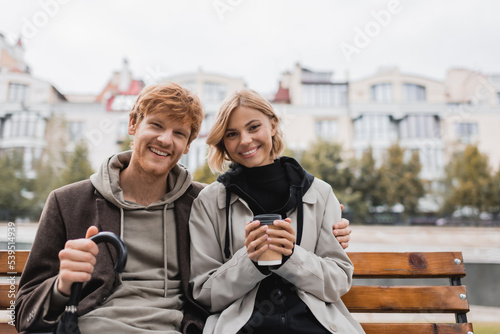happy young man with umbrella hugging girlfriend holding paper cup while sitting on bench.