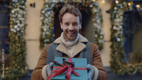 Portrait of the happy smiled caucasian man standing on the decorated street wearing earphones holding and showing the green christmas gift with with red ribbon looking at the camera