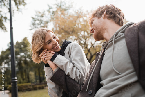 joyful and blonde woman holding hand of redhead boyfriend while smiling in park.