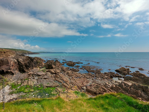 The rocky coast of the Celtic Sea on a sunny day. Clear blue sky with white clouds over the seascape. Green grass on the shore.
