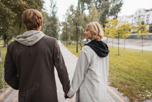joyful blonde woman looking at cheerful man while holding hands and strolling in park.