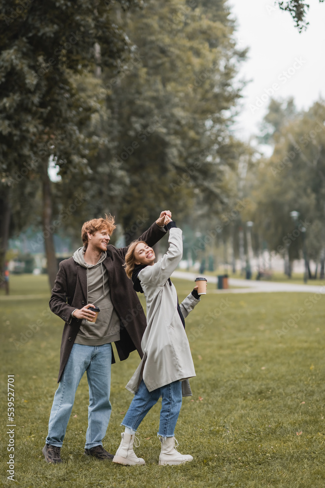 full length of happy redhead man and cheerful woman holding coffee to go while twirling during dance in park.