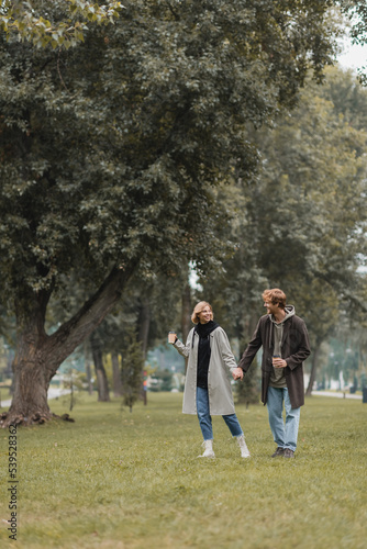 full length of positive redhead man and cheerful woman holding coffee to go while walking in park.