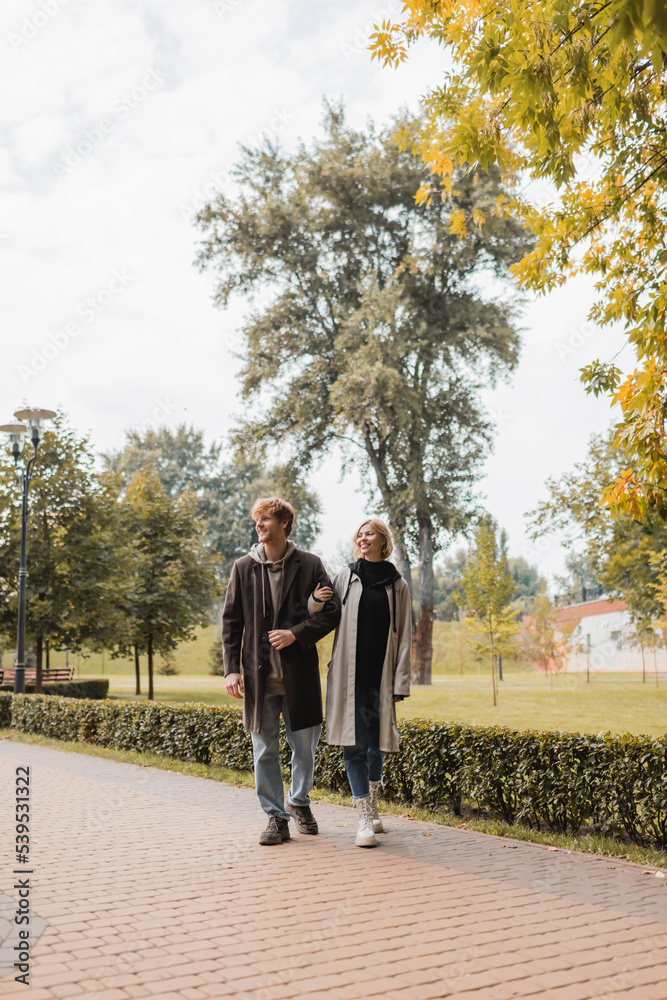 full length of young and positive couple in coats walking together in autumnal park during date. full length of young and positive couple in coats walking together in autumnal park during date.