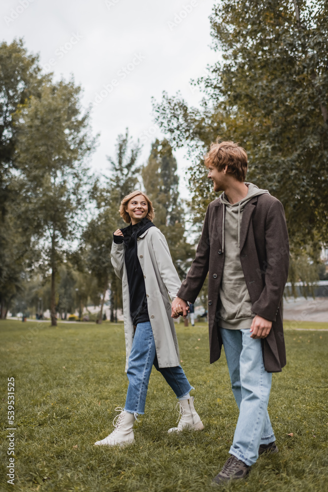 full length of young man in coat holding hands with cheerful girlfriend while walking in autumnal park.