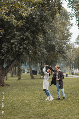 full length of happy redhead man and cheerful woman holding coffee to go while dancing in park.