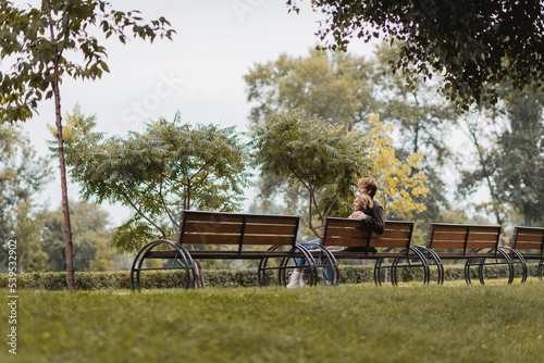 cheerful young man and woman hugging and sitting on wooden bench in green park.