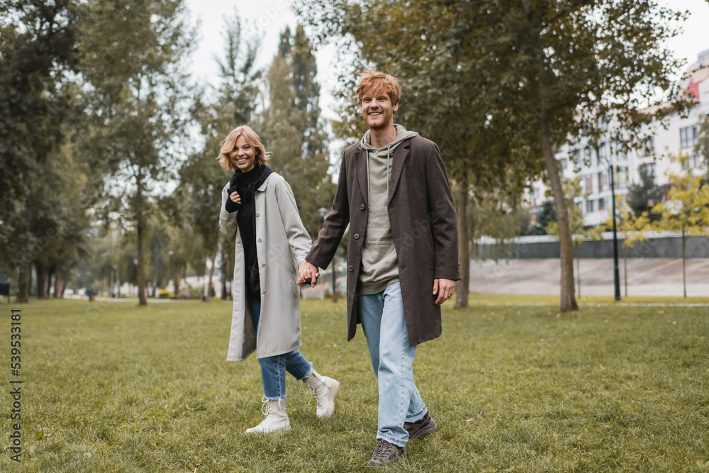 full length of happy young man in coat holding hands with cheerful girlfriend while walking in autumnal park. full length of happy young man in coat holding hands with cheerful girlfriend while walking in autumnal park.