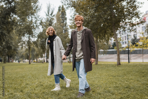full length of happy young man in coat holding hands with cheerful girlfriend while walking in autumnal park.