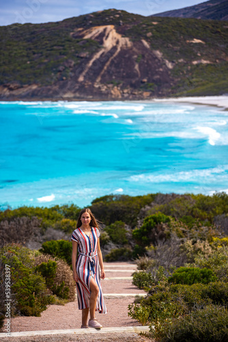 a beautiful long-haired girl in a long dress walks along a path overlooking a paradise beach with turquoise water and mountains; panorama of hellfire bay in western australia seen from above