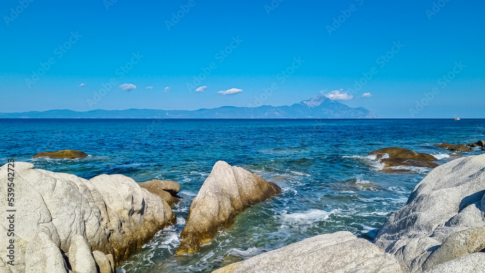 Naklejka premium Beautiful panoramic view of holy Mount Athos seen from Karydi beach, peninsula Sithonia, Chalkidiki (Halkidiki), Greece, Europe. Summer vacation at Aegean Mediterranean Sea. Rock formations in the bay