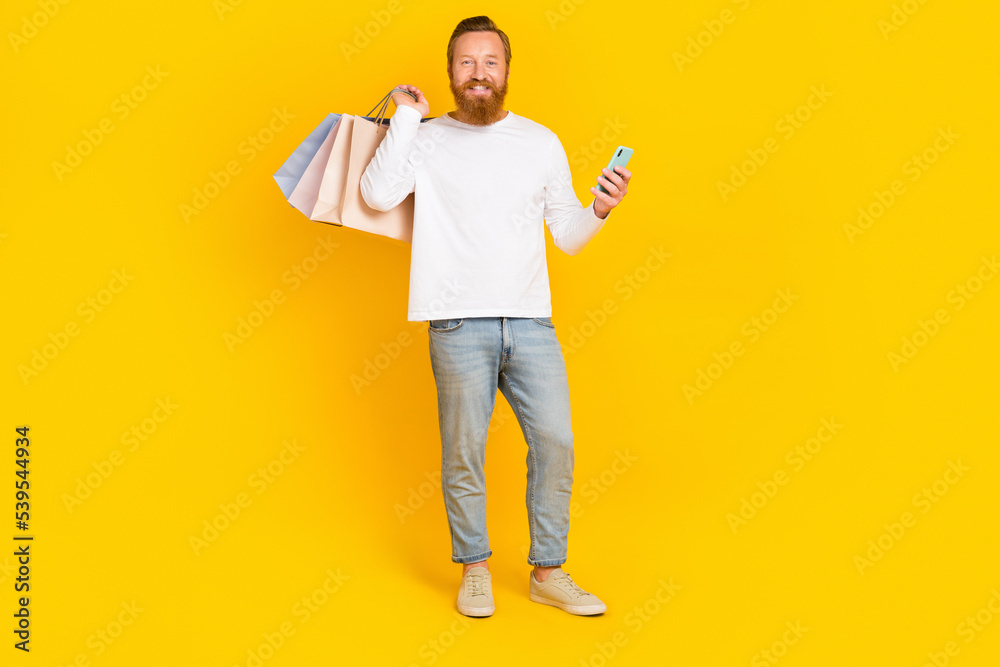 Full length photo of funky beard man hold bags telephone wear white shirt jeans boots isolated on yellow color background Full length photo of funky beard man hold bags telephone wear white shirt jeans boots isolated on yellow color background