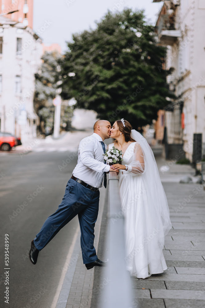 A stylish adult groom and a beautiful smiling bride in a white dress are hugging, kissing on the street in the city near the railings. Wedding photography, portrait.