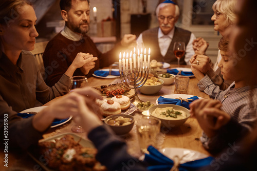 Close up of extended family holding hands and praying while celebrating Hanukkah at dining table.