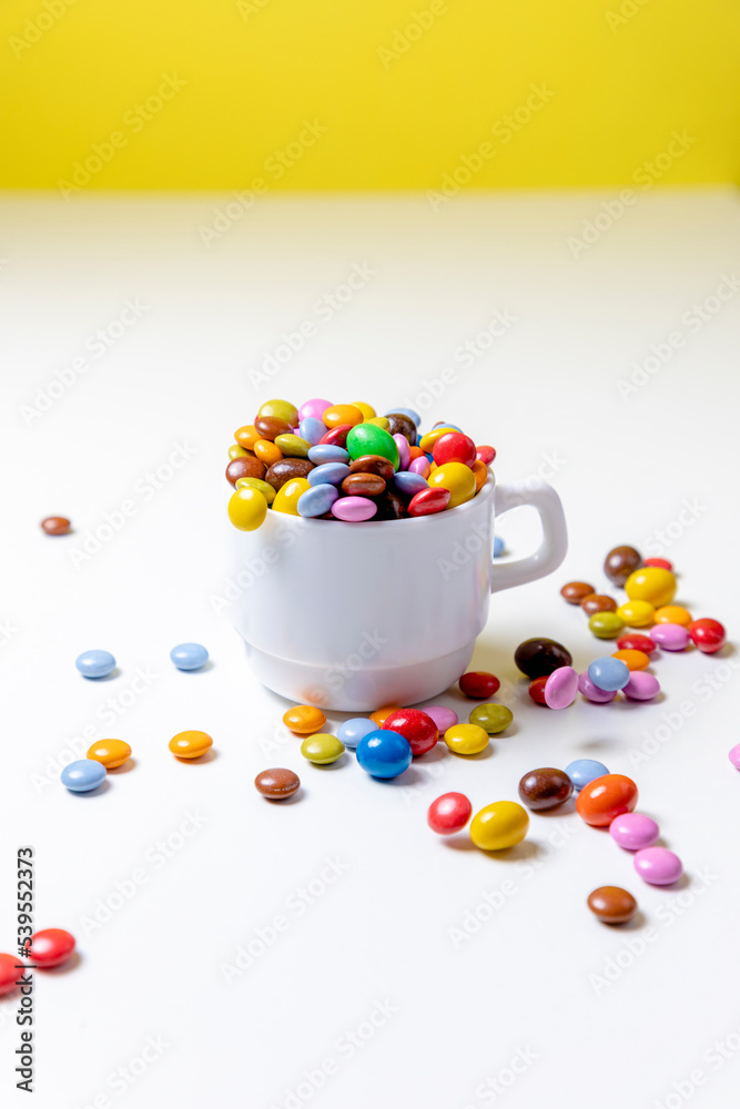 Taza blanco con bolas de chocolate de colores con fondo blanco y amarillo y mano cogiendo golosinas