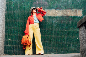 Hipster young woman in bright clothes, sun glasses, backpack bag and bucket hat posing on the green tile wall background. Urban city street fashion. Fashion blogger. Selective focus. Copy space