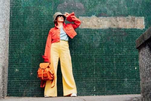 Hipster young woman in bright clothes, sun glasses, backpack bag and bucket hat posing on the green tile wall background. Urban city street fashion. Fashion blogger. Selective focus. Copy space