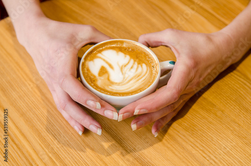 Young female hands holding and hugging a cup of coffee with latte art at a wooden table in a cafe