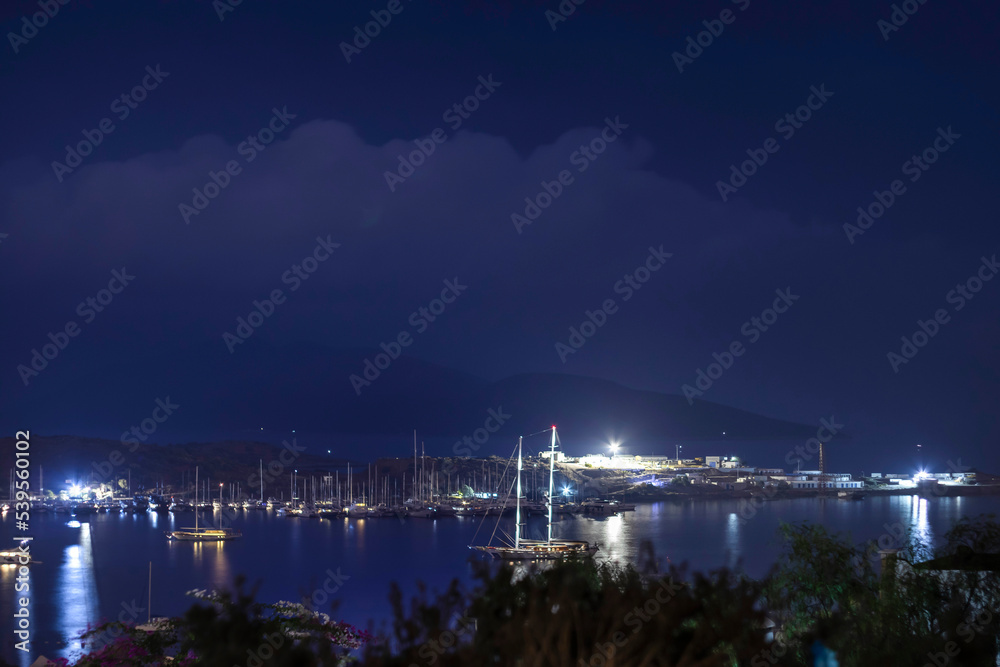 Fototapeta premium Boat on the sea, harbor and lights reflecting on the sea at blue hour on a rainy and cloudy evening.