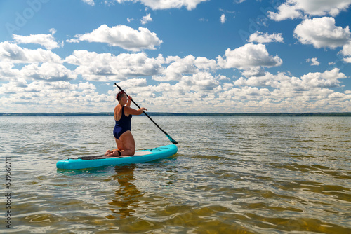 a woman in a closed swimsuit with an informal hairstyle on her knees on a SUP board with a paddle floats on the water.