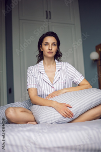 portrait of a caucasian woman sitting on a bed in striped pajamas with a pillow at home in the morning and enjoying a new day. © saulich84