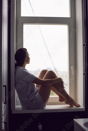 woman in striped pajamas sits on the windowsill in the bathroom in the morning and thinks about happiness.
