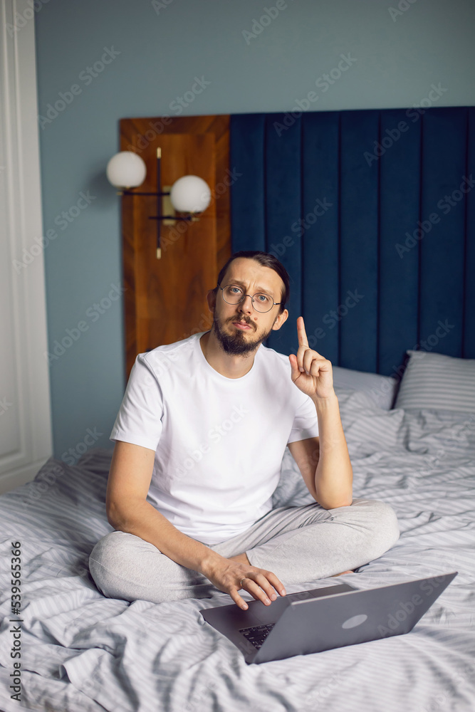 Fototapeta premium man in a white T-shirt, a freelancer with glasses, sits on the bed at home with a laptop and works remotely in the morning. suddenly came a brilliant idea