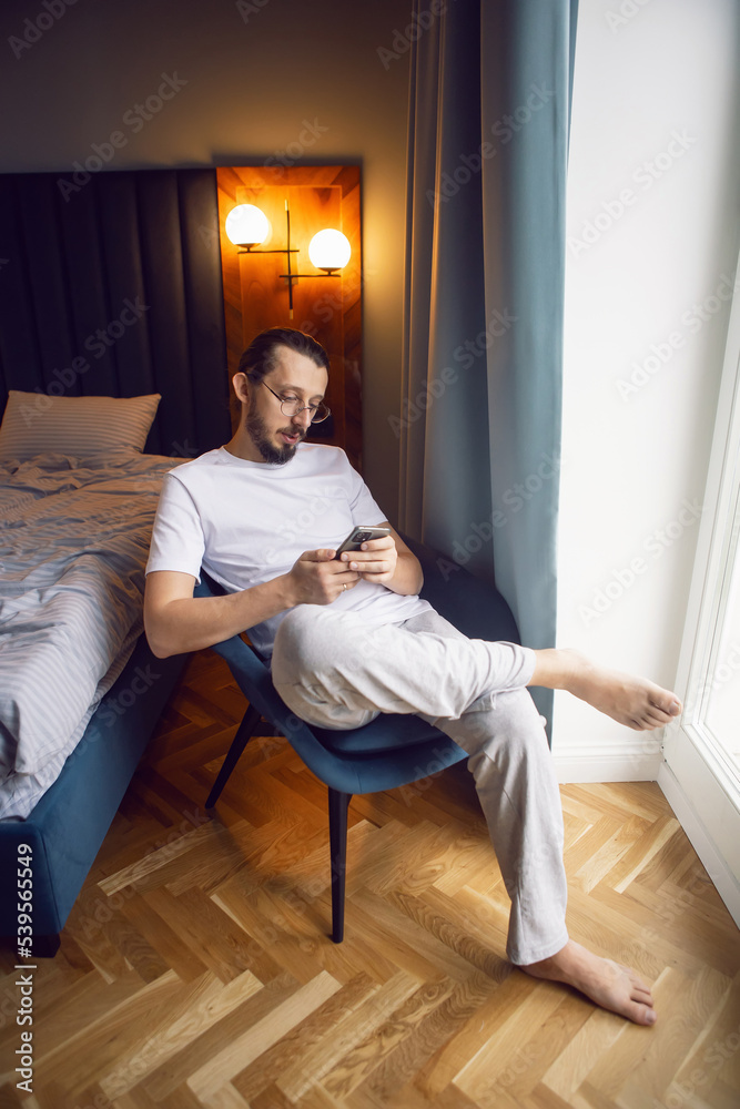 portrait of a bearded man in glasses, white T-shirt and house pants sitting on a chair by the window with a phone. by the bed.