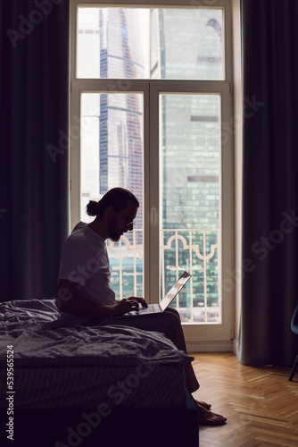 man in a white T-shirt, a freelancer with glasses, sits on the bed at home with a laptop and works remotely in the morning. in front of a large window.