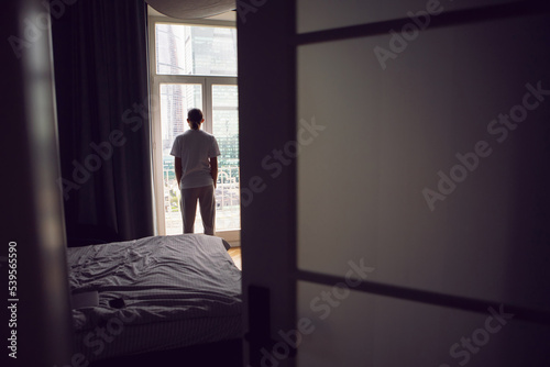 man with long hair, beard and glasses stands at a large window in an apartment with a view of a skyscraper in the morning. the guy is dressed in a white T-shirt and sweatpants barefoot.