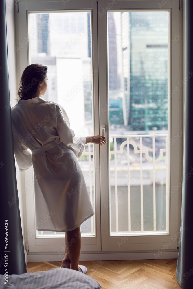 portrait of a beautiful brunette woman in a bathrobe stands at a large window in an apartment opposite a skyscraper in the morning. - obrazy, fototapety, plakaty 