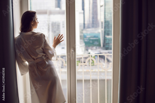 portrait of a beautiful brunette woman in a bathrobe stands at a large window in an apartment opposite a skyscraper in the morning.