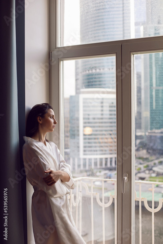 portrait of a beautiful brunette woman in a bathrobe stands at a large window in an apartment opposite a skyscraper in the morning. Free Download portrait of a beautiful brunette woman in a bathrobe stands at a large window in an apartment opposite a skyscraper in the morning. - GFXLoad