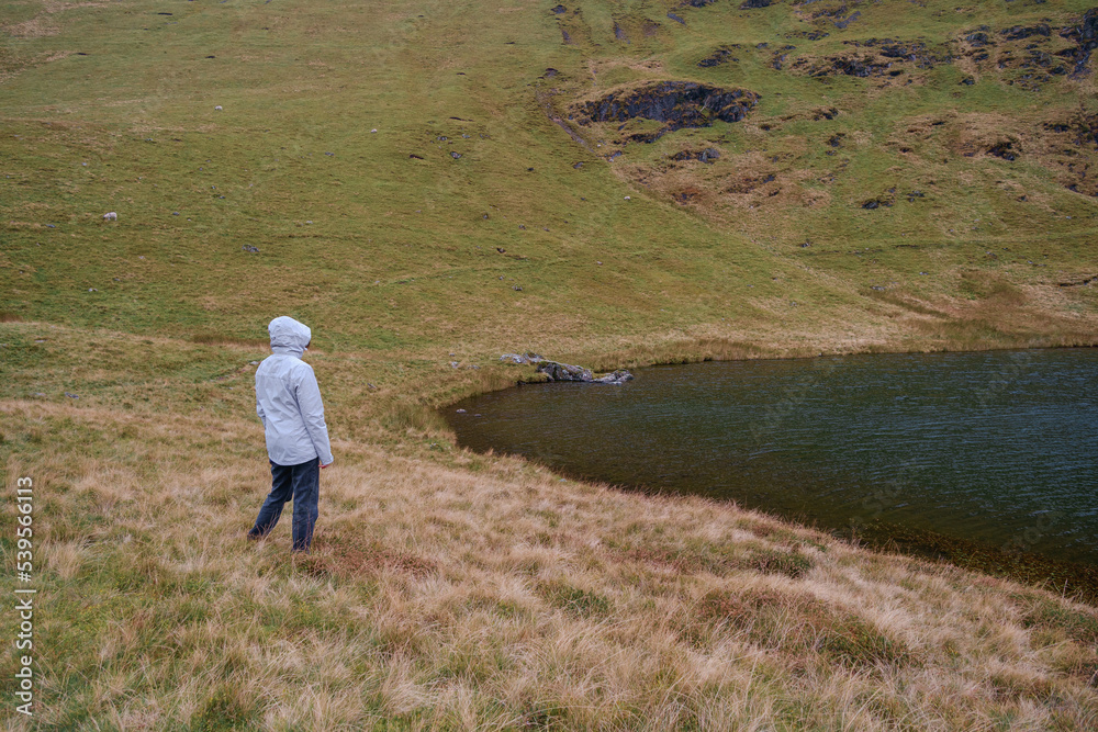 Person walking or hiking in raincoat near to Bowscale Tarn lake in mountains, United Kingdom Person walking or hiking in raincoat near to Bowscale Tarn lake in mountains, United Kingdom