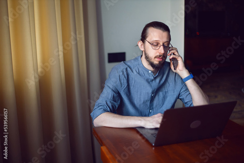 portrait of a businessman a man with a beard and glasses is sitting at a laptop at a wooden table at home. the freelancer works remotely next to the window the light is natural. talking on the phone.