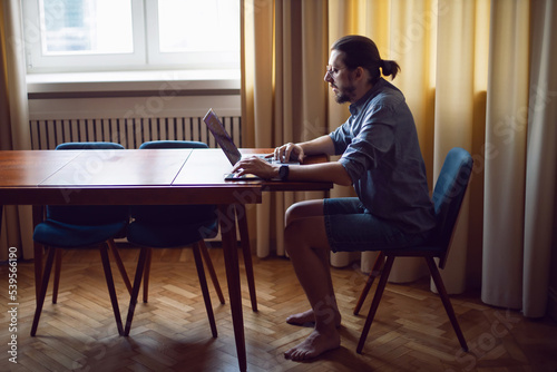 portrait of a businessman a man with a beard and glasses is sitting at a laptop at a wooden table at home. the freelancer works remotely next to the window the light is natural.