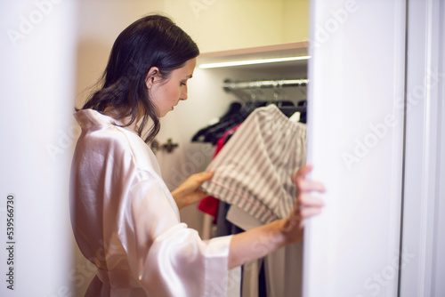 woman in a white bathrobe stands at the wardrobe with clothes and chooses a shirt.