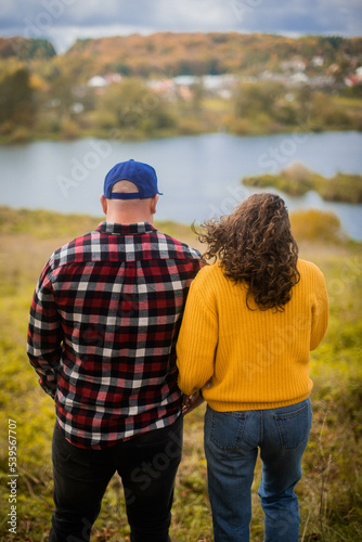 curly-haired woman holding her husband's hand. the woman is wearing a yellow sweater and blue jeans. the man is wearing a checkered shirt, black jeans and a blue cap. looking at the autumn river