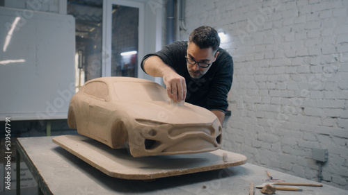 Senior automotive designer sculpts the prototype model of the car made of clay using sculpting tools. Experienced designer works in a workshop. Automotie industry.