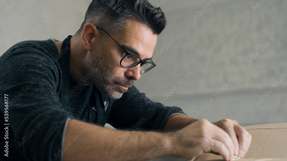 Experienced ceramist with an apron uses sculpting tool with both hands to create the design details on the sculpture of a prototype, eco friendly model of a car made of raw clay.