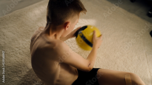 Young enthusiastic boy training abs holds a football while exercising alone indoors. Prepares before the soccer match. Sport and active lifestyle concept.