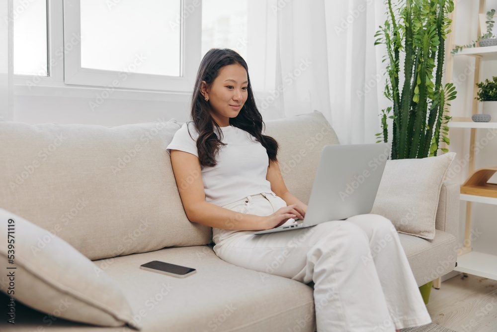Young Asian woman looking at laptop screen on her lap sitting on couch at home lifestyle work as a freelancer Young Asian woman looking at laptop screen on her lap sitting on couch at home lifestyle work as a freelancer