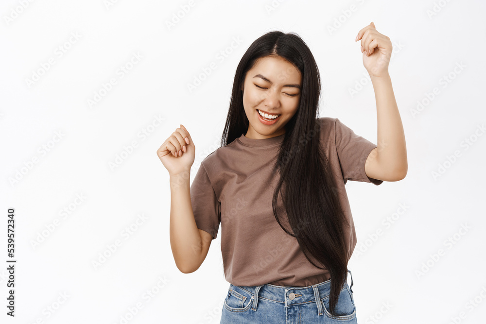Happy dancing asian girl, celebrating and triumphing, winning and cheering, standing over white studio background