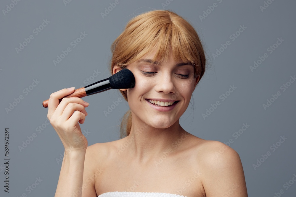 a happy, joyful woman stands on a gray background and paints her face with a fluffy makeup brush, closing her eyes with pleasure a happy, joyful woman stands on a gray background and paints her face with a fluffy makeup brush, closing her eyes with pleasure