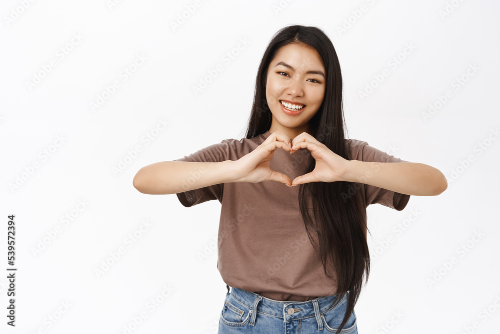 Cute young asian woman shows heart, love sign, express care and tenderness, stands in tshirt over white background Cute young asian woman shows heart, love sign, express care and tenderness, stands in tshirt over white background