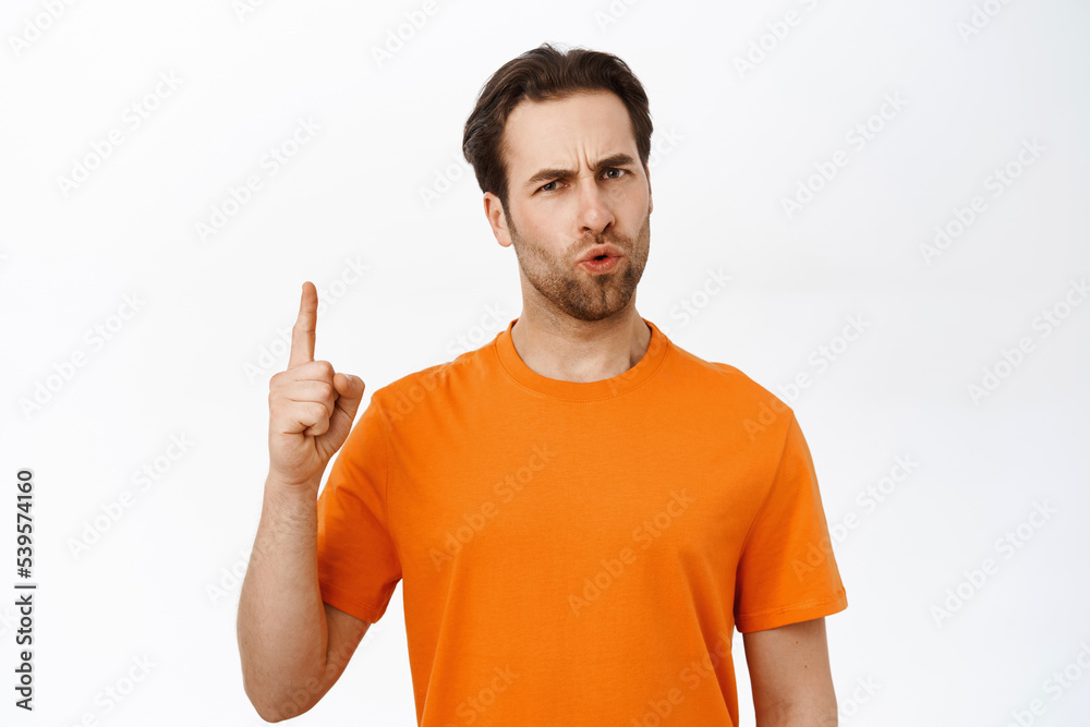 Portrait of disappointed guy pointing finger up, grimacing, being upset, standing in orange tshirt