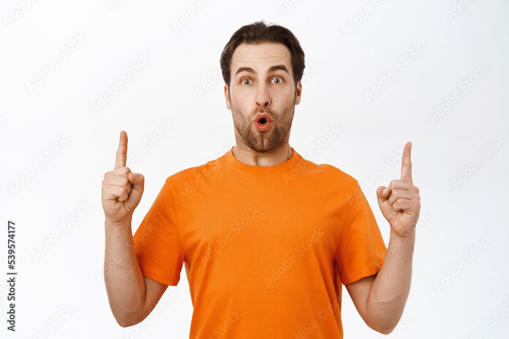 Impressed man points up, shows smth on top, making impressed face expression, standing over white background