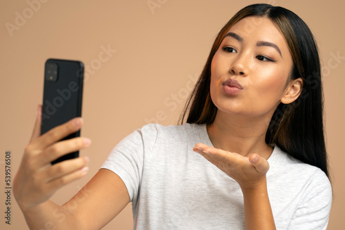 Joyful asian young woman using smartphone for video call sending air kiss to webcam