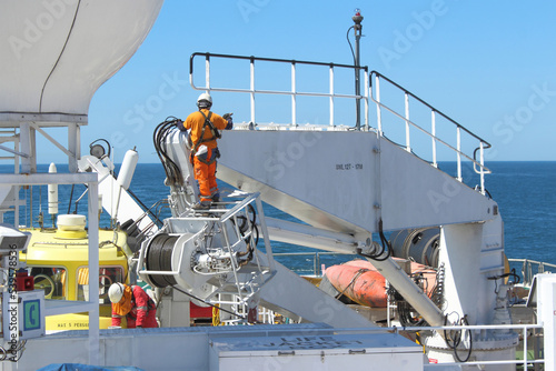 Two seafarers painting ship\'s crane while out at sea. Both wearing full PPE and fall protection harnesses to prevent fall and injury. Onboard safety culture concept.