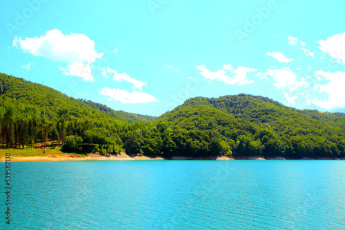 Heavenly view at Gerosa Lake with a cyan sky and some white fluffy clouds suspended above a bulky mass of green vegetation sitting on rough hills and some clear azure waters further below